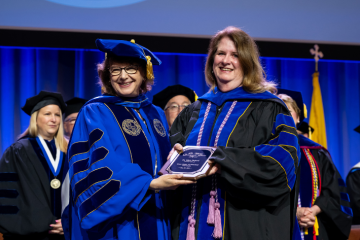 Mary Packard wearing regalia receives award plaque from President Yam